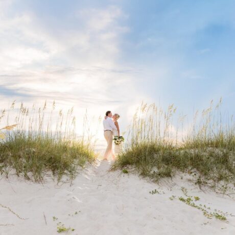A couple stands together on a sandy beach dune surrounded by tall grass, with the sun shining through a partly cloudy sky. The person in front holds a bouquet of flowers.