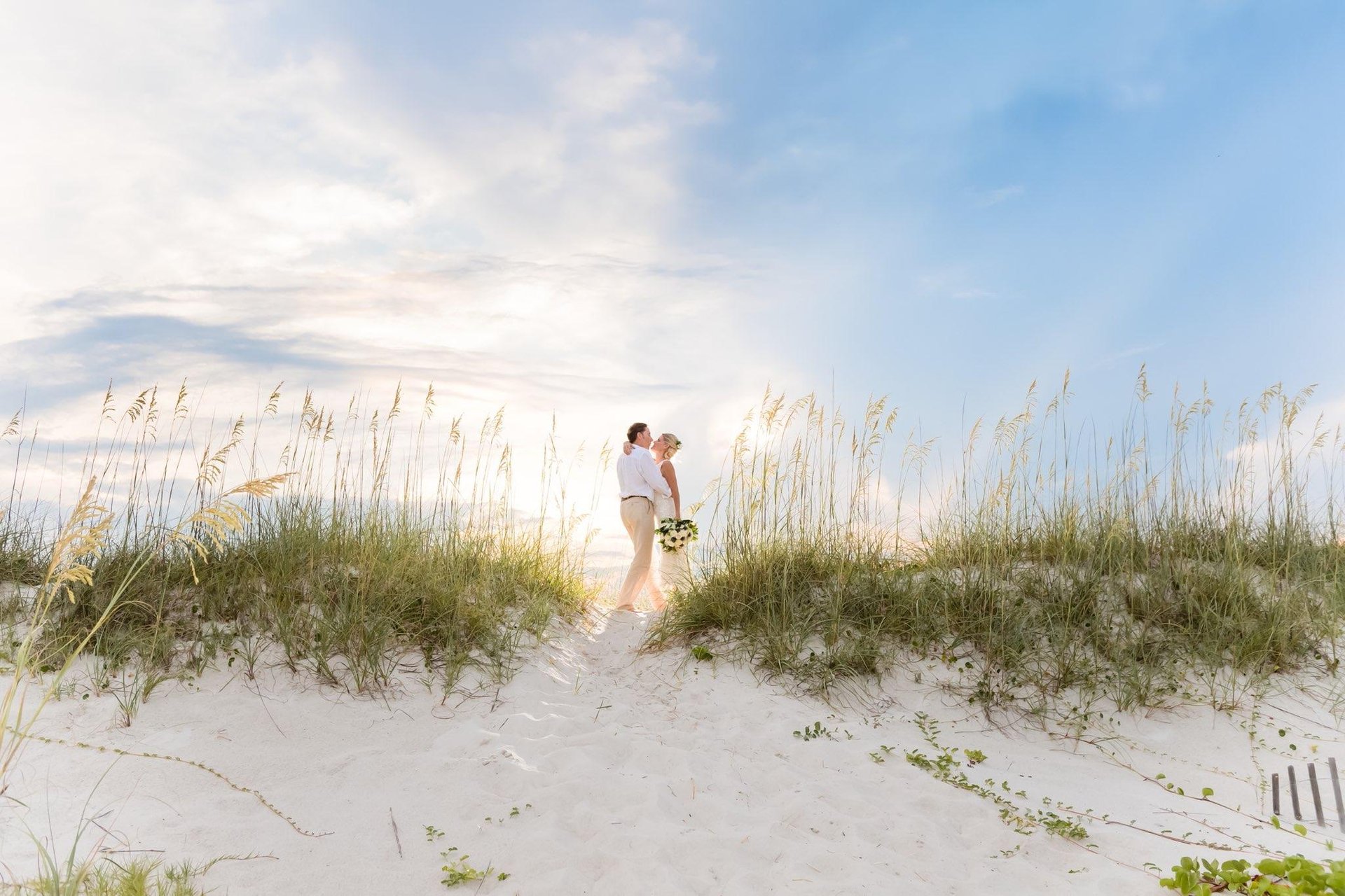 A couple stands together on a sandy beach dune surrounded by tall grass, with the sun shining through a partly cloudy sky. The person in front holds a bouquet of flowers.