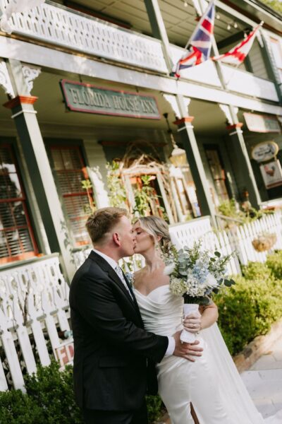 A bride and groom share a kiss outside the Florida House Inn, surrounded by greenery and white fencing. The bride holds a bouquet, and both are dressed in formal wedding attire. Flags hang above the porch behind them.