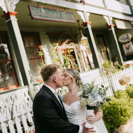 A bride and groom share a kiss outside the Florida House Inn, surrounded by greenery and white fencing. The bride holds a bouquet, and both are dressed in formal wedding attire. Flags hang above the porch behind them.