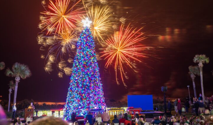 A large, brightly lit Christmas tree with a star topper stands surrounded by a crowd at night. Colorful fireworks explode in the sky above, and palm trees are visible in the background.