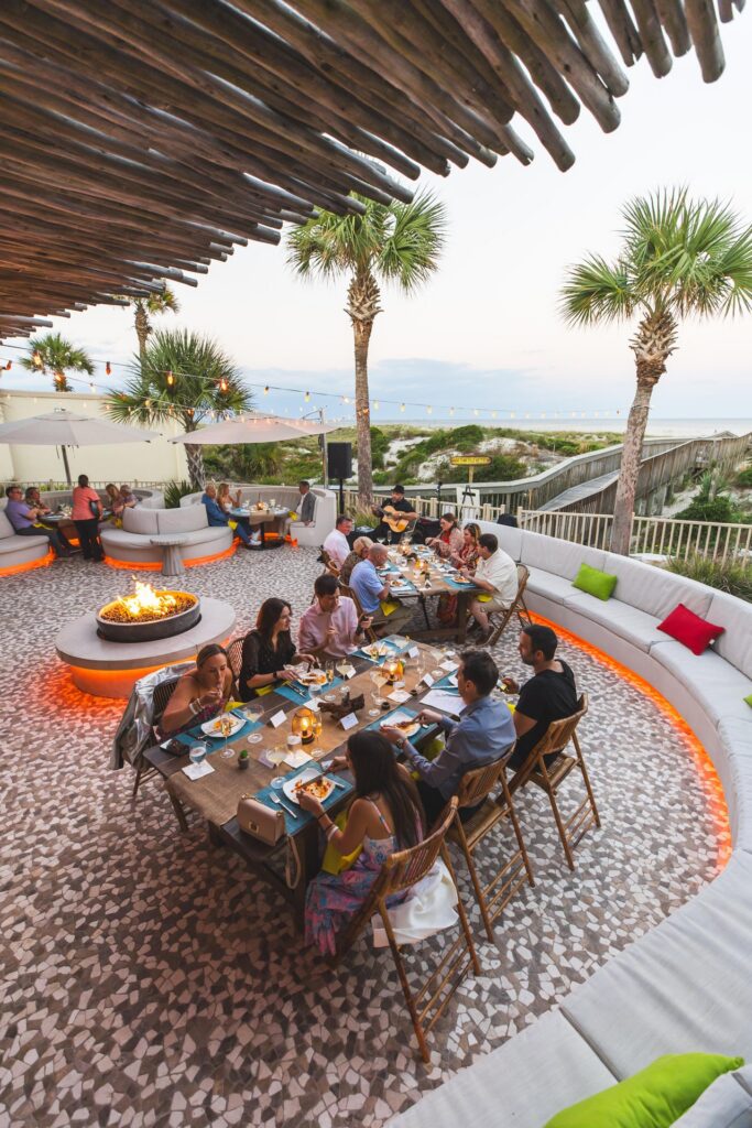 A group of people are dining at a long outdoor table on a patio by the beach, surrounded by palm trees, string lights, a fire pit, and curved cushioned seating at sunset.