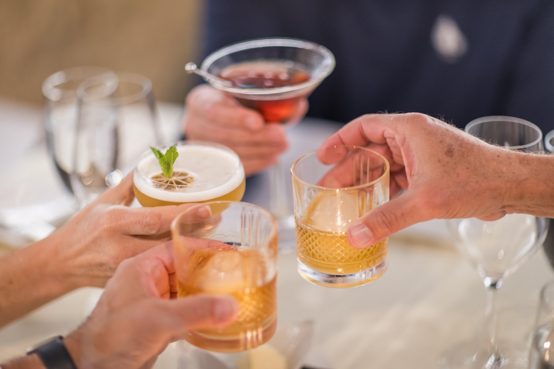 Four people clink glasses with various cocktails over a table, celebrating together. The drinks include whiskey, a cocktail with foam and a lime slice, and a martini. The setting appears casual and festive.