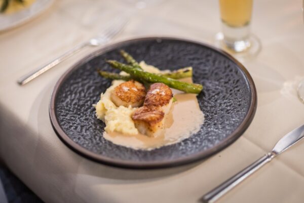A dark plate with seared scallops on creamy mashed potatoes, topped with light sauce and served with grilled asparagus, set on a cream-colored tablecloth with a knife, fork, and a glass in the background.