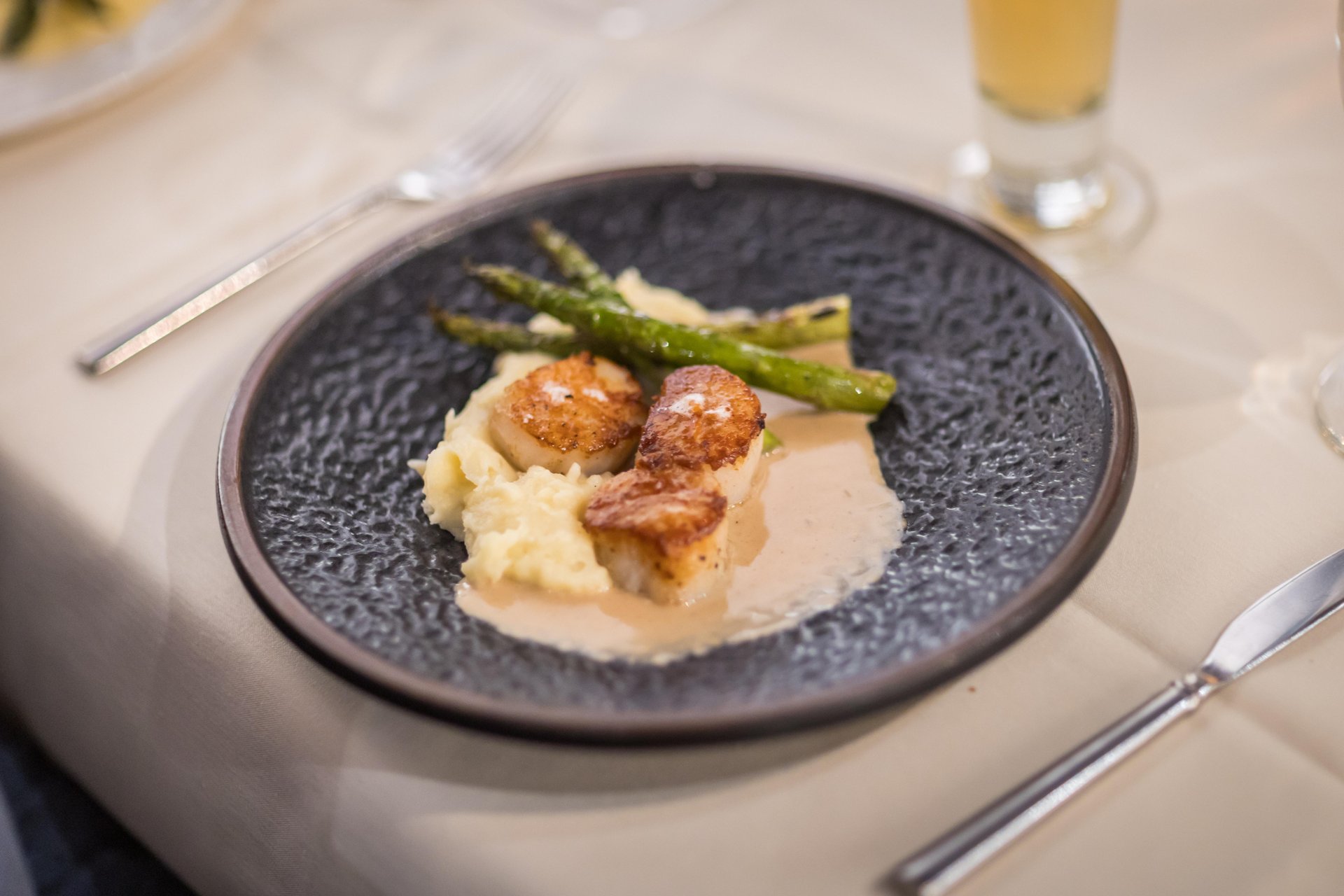 A dark plate with seared scallops on creamy mashed potatoes, topped with light sauce and served with grilled asparagus, set on a cream-colored tablecloth with a knife, fork, and a glass in the background.