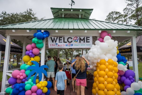 People gather under a gazebo decorated with colorful balloons and a sign reading Welcome Island Hop Craft Beer Fest. Palm tree and peace symbols are part of the decor, and trees and tents are visible in the background.