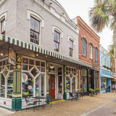 Street view of colorful shops and cafes with large windows and striped awnings on a tree-lined sidewalk with outdoor seating and people relaxing under palm trees.
