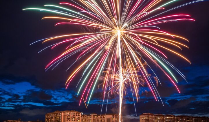 Colorful fireworks burst over a beach at night, illuminating the sky and reflecting on the water, with several buildings and a dark blue, cloudy sky in the background.