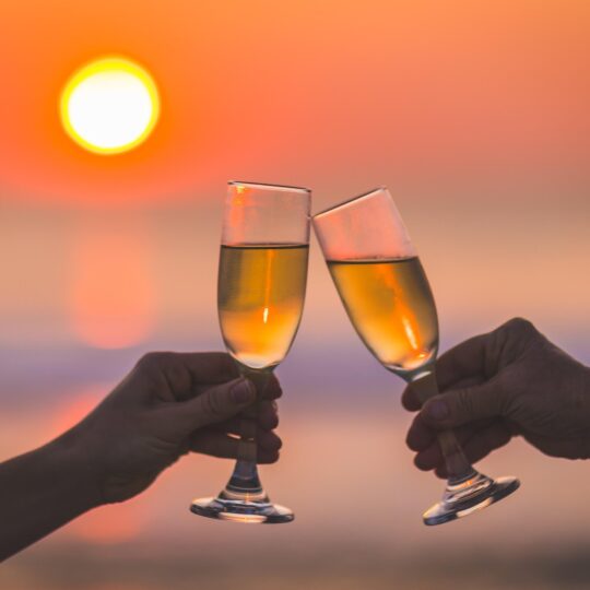 Two hands clink champagne glasses together with a vibrant orange sunset over the ocean in the background, celebrating or toasting at the beach.