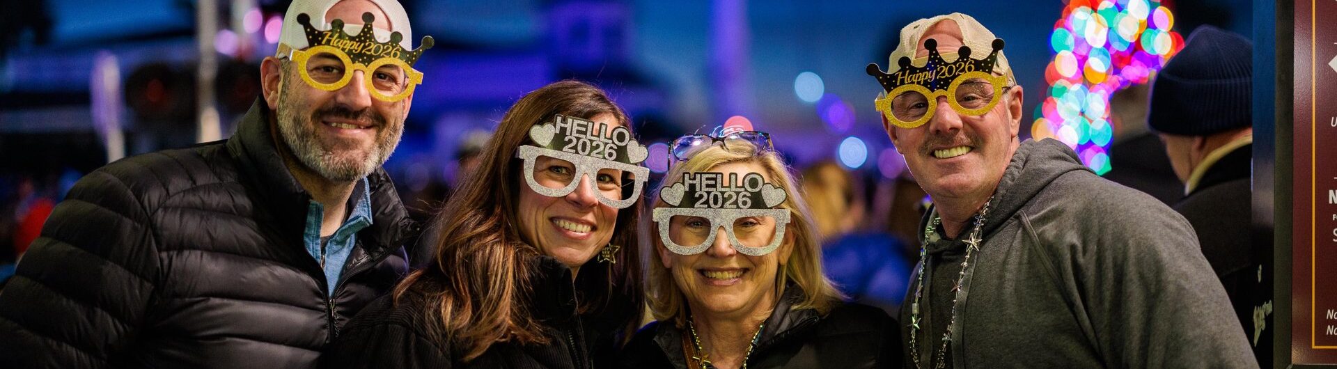 Four people wearing festive Hello 2024 glasses and crowns smile together outdoors at night. They are dressed warmly, and colorful holiday lights and a Christmas tree appear in the blurred background.