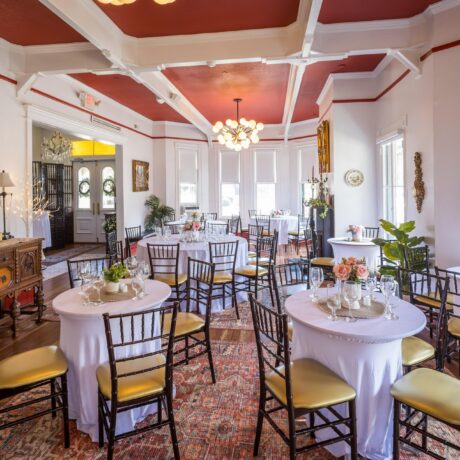 Bright, elegant dining room with round tables covered in white cloths, gold-cushioned chairs, floral centerpieces, wooden sideboard, large windows, and red ceiling with white beams. Natural light fills the room.