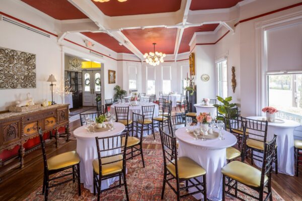 Bright, elegant dining room with round tables covered in white cloths, gold-cushioned chairs, floral centerpieces, wooden sideboard, large windows, and red ceiling with white beams. Natural light fills the room.