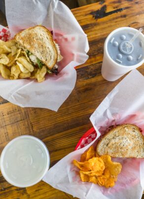 Two baskets with sandwiches and chips sit on a wooden table, each accompanied by a paper cup with a lid and straw. One basket has potato chips; the other has orange chips, possibly barbecue-flavored.