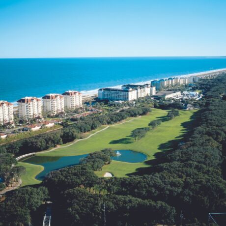 Aerial view of a coastal resort area with tall beachfront buildings, a golf course with a pond, and the ocean stretching along the horizon under a clear blue sky.