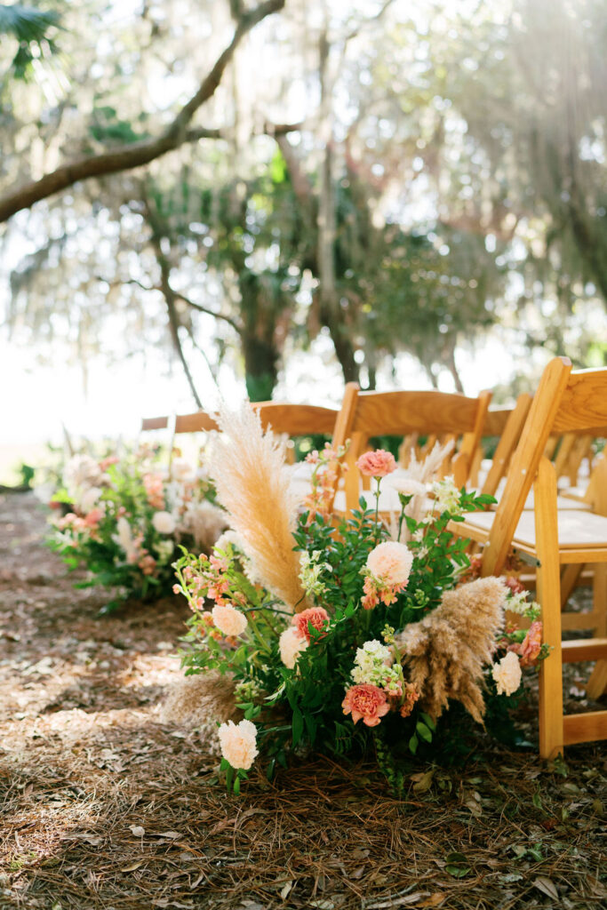 Rows of wooden chairs are set up outdoors on pine needles, with lush floral arrangements featuring pampas grass and pink, peach, and white flowers. Sunlight filters through moss-draped trees in the background.