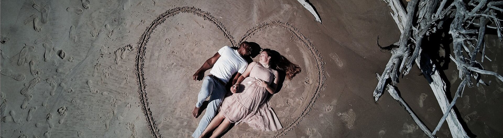 A couple lies side by side on sandy ground inside a large heart shape drawn in the sand, holding hands near driftwood.
