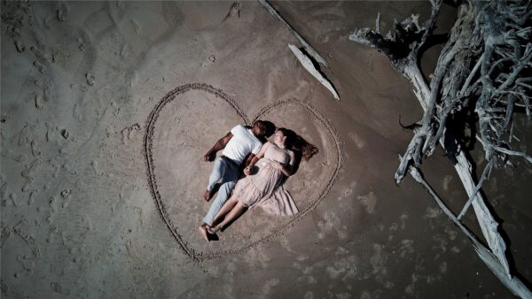 A couple lies side by side on sandy ground inside a large heart shape drawn in the sand, holding hands near driftwood.