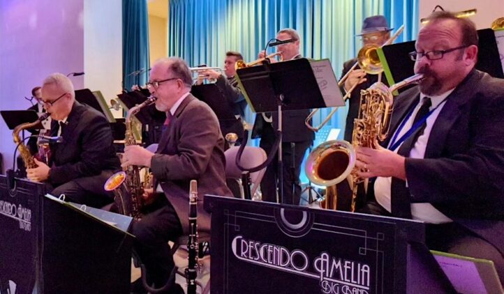 A jazz big band performs on stage, with musicians playing saxophones, trombones, and trumpets. They sit behind music stands labeled Crescendo Amelia Big Band against a blue curtain backdrop.