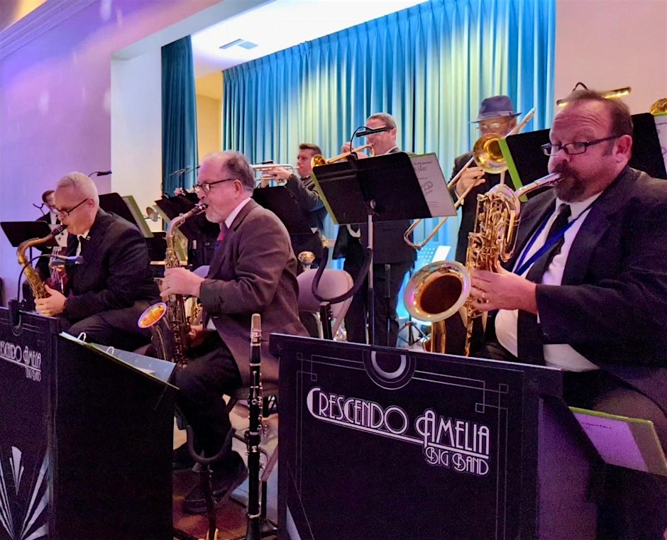A jazz big band performs on stage, with musicians playing saxophones, trombones, and trumpets. They sit behind music stands labeled Crescendo Amelia Big Band against a blue curtain backdrop.