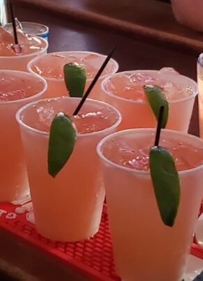 Eight plastic cups filled with a light orange drink, each garnished with a lime wedge and a black straw, are lined up on a bar mat. A tall glass with a logo and a straw is beside them.