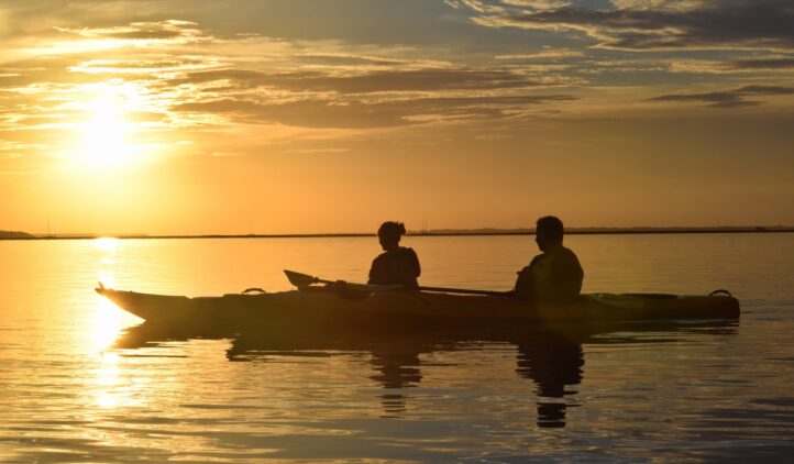 Two people in a kayak paddle on calm water at sunset, silhouetted against a golden sky with scattered clouds. The sunlight reflects off the water, creating a peaceful and serene scene.