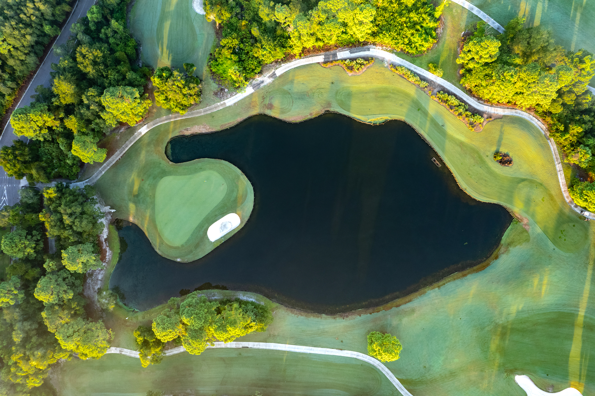 Aerial view of a golf course with a green island surrounded by a dark pond, winding paths, and lush green trees and fairways bathed in sunlight.