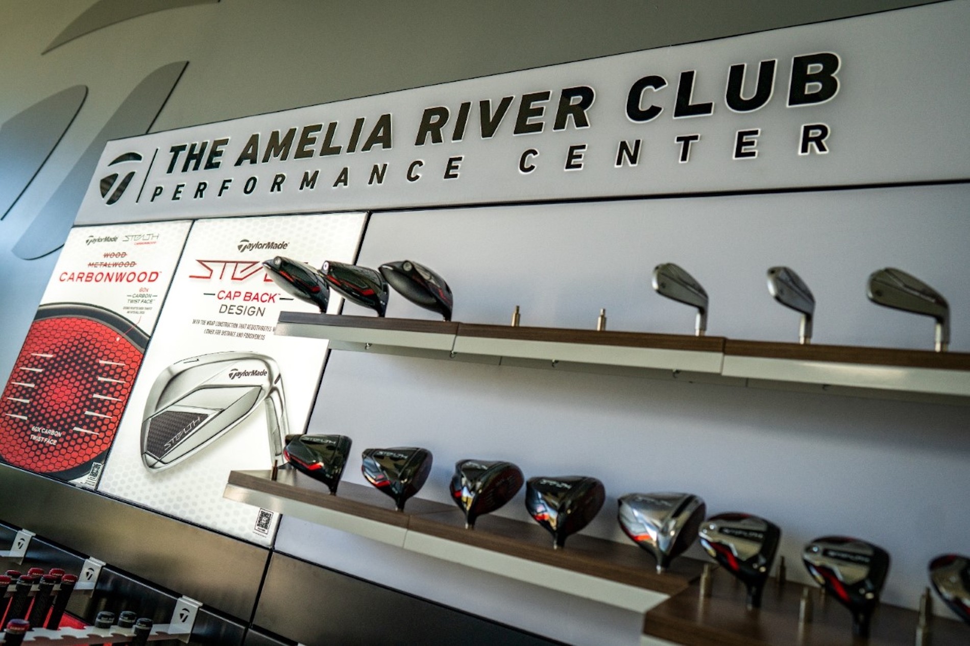 A display wall at The Amelia River Club Performance Center showcases various TaylorMade golf club heads and equipment, neatly arranged on shelves with branded signage above.