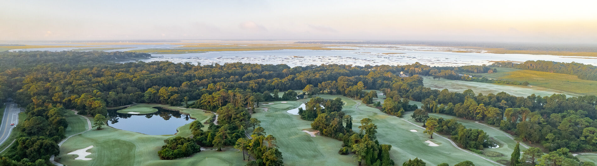 Aerial view of a lush, green golf course surrounded by trees and small ponds, with a road on the left and a distant body of water under a clear blue sky at sunrise.