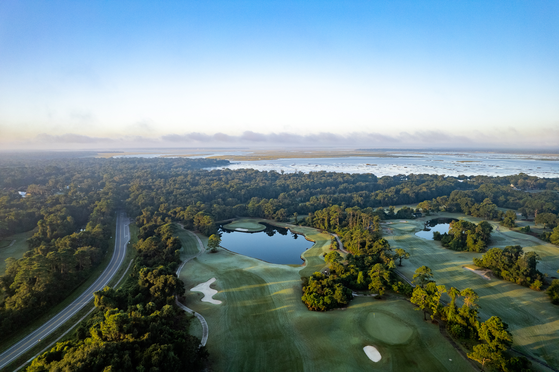 Aerial view of a golf course with green fairways, a small lake, surrounding trees, a road to the left, and a distant marshland under a clear blue sky.