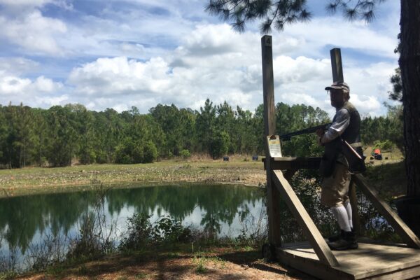 A person stands on a wooden shooting platform holding a shotgun, overlooking a pond and surrounded by trees under a partly cloudy sky.