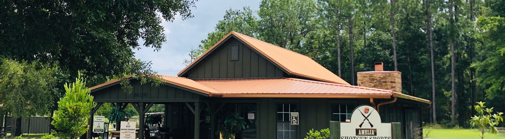 A green building with a brown metal roof stands surrounded by trees and landscaping. A sign in front reads Shotgun Sports Center. The sky is partly cloudy above the rural setting.