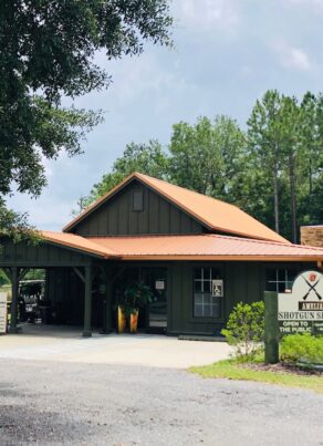 A green building with a brown metal roof stands surrounded by trees and landscaping. A sign in front reads Shotgun Sports Center. The sky is partly cloudy above the rural setting.