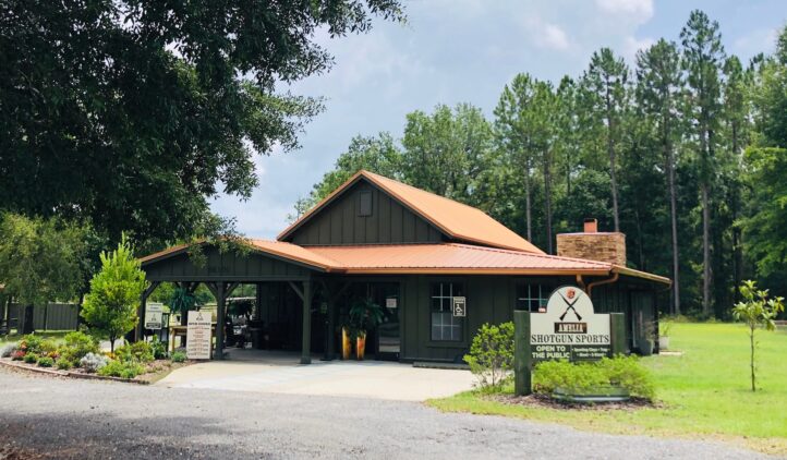 A green building with a brown metal roof stands surrounded by trees and landscaping. A sign in front reads Shotgun Sports Center. The sky is partly cloudy above the rural setting.