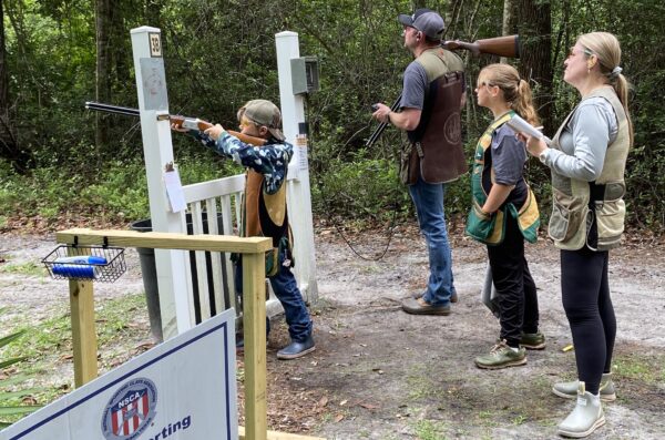 Four people stand at an outdoor shooting range. A boy aims a shotgun at a target, while two women and a man observe. All wear safety vests and glasses, surrounded by trees and shooting equipment.