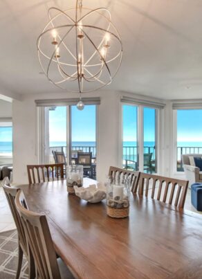 A spacious, modern dining and living area with large windows showcasing an ocean view, wooden dining table with chairs, cozy seating, built-in shelves, and abundant natural light.