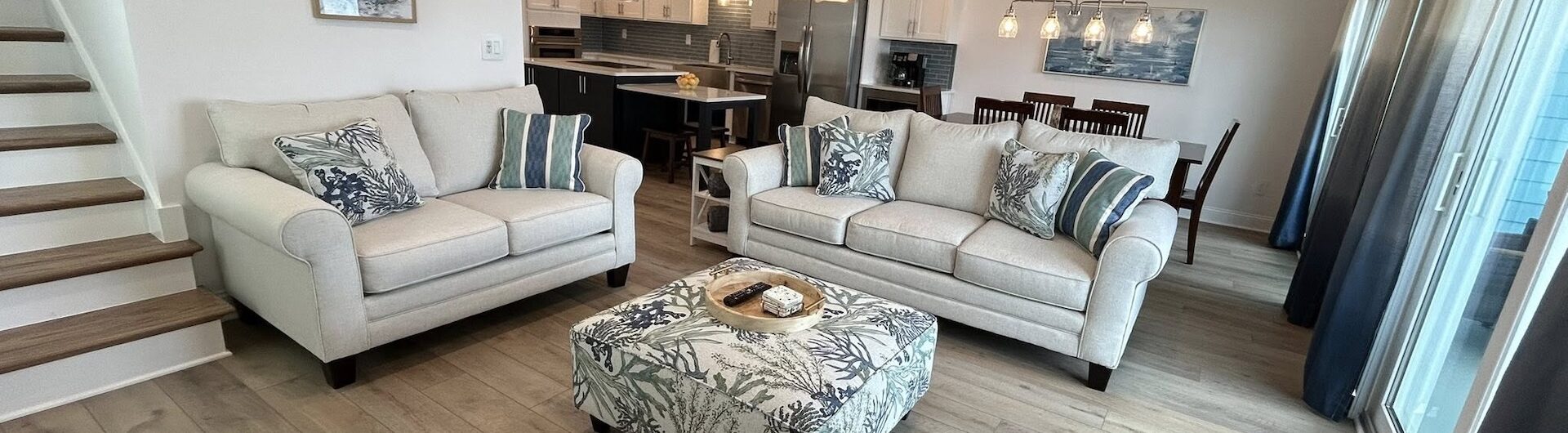 Modern living room with light wood flooring, beige sofas with striped pillows, a patterned ottoman, navy chair, open kitchen, dining area, staircase, and large windows letting in natural light.