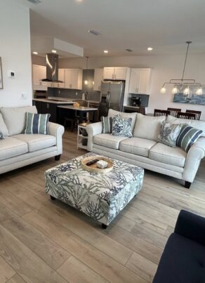 Modern living room with light wood flooring, beige sofas with striped pillows, a patterned ottoman, navy chair, open kitchen, dining area, staircase, and large windows letting in natural light.