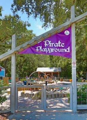 Entrance to Pirate Playground, marked by a purple sign, with trees, plants, and playground equipment visible inside the fenced area under a sunny sky.