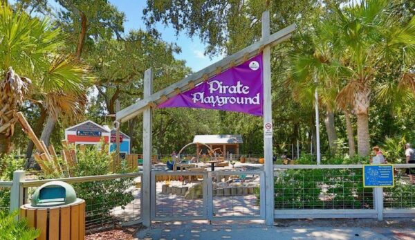 Entrance to Pirate Playground, marked by a purple sign, with trees, plants, and playground equipment visible inside the fenced area under a sunny sky.