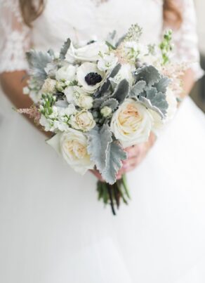 A bride in a white lace wedding dress holds a bouquet of white and cream flowers, including roses, anemones, and dusty miller leaves, with soft green and pink accents.