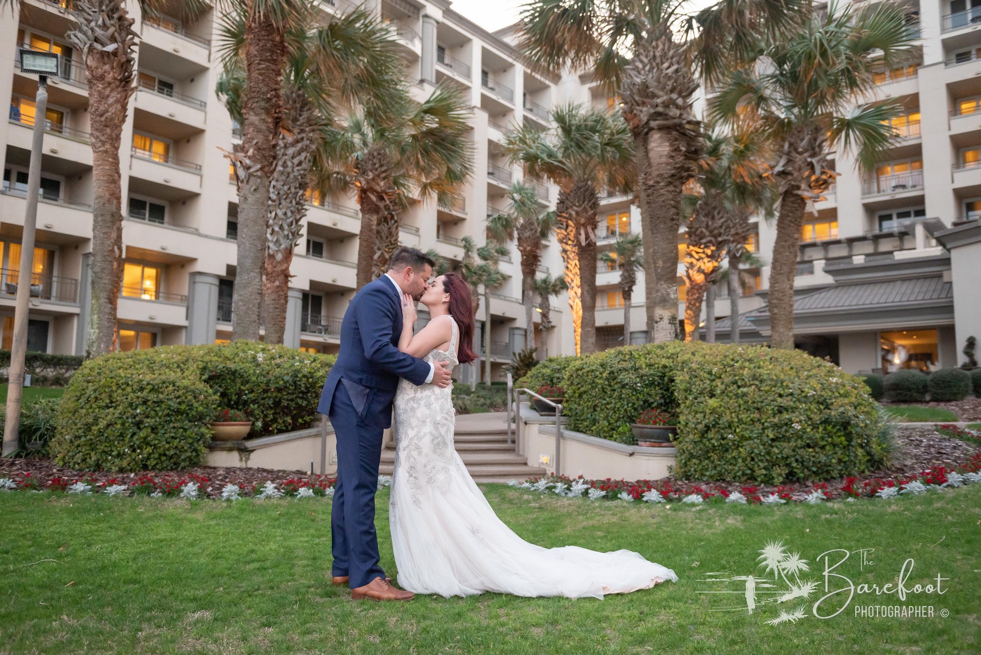 A bride and groom share a kiss on a grassy lawn, surrounded by palm trees and flower beds, with a multi-story hotel building in the background at sunset.