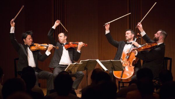 Four male musicians in tuxedos play string instruments on stage, raising their bows dramatically in unison, with sheet music in front of them and an audience in the foreground.