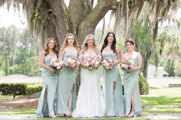 Five women in light green dresses and one in a white gown stand outdoors in front of a large tree, holding pink and white flower bouquets, smiling for a wedding photo.