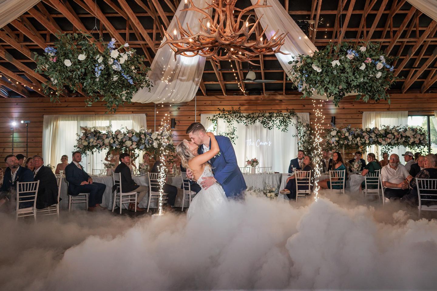 A bride and groom share their first dance in a rustic, decorated venue with wooden beams, floral arrangements, draped fabric, and artificial fog on the floor, surrounded by seated guests watching them.