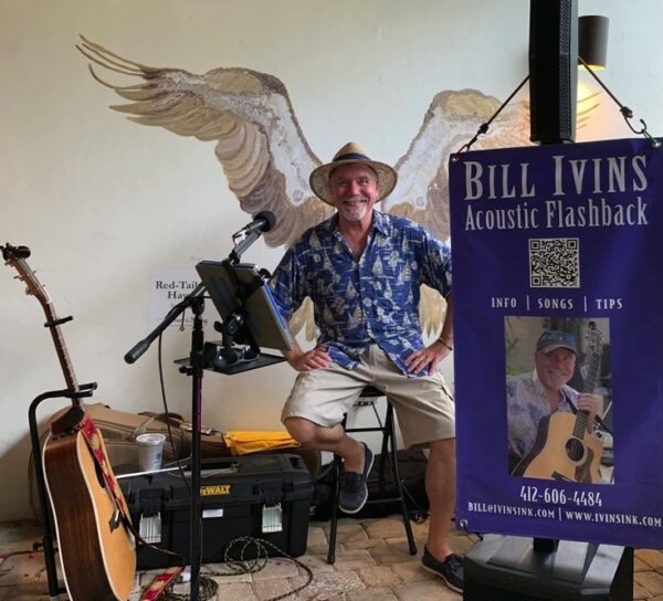 A smiling man in a blue shirt and straw hat sits in front of a microphone and music setup, with a guitar beside him and painted bird wings on the wall behind. A banner for Bill Ivins Acoustic Flashback is displayed on the right.