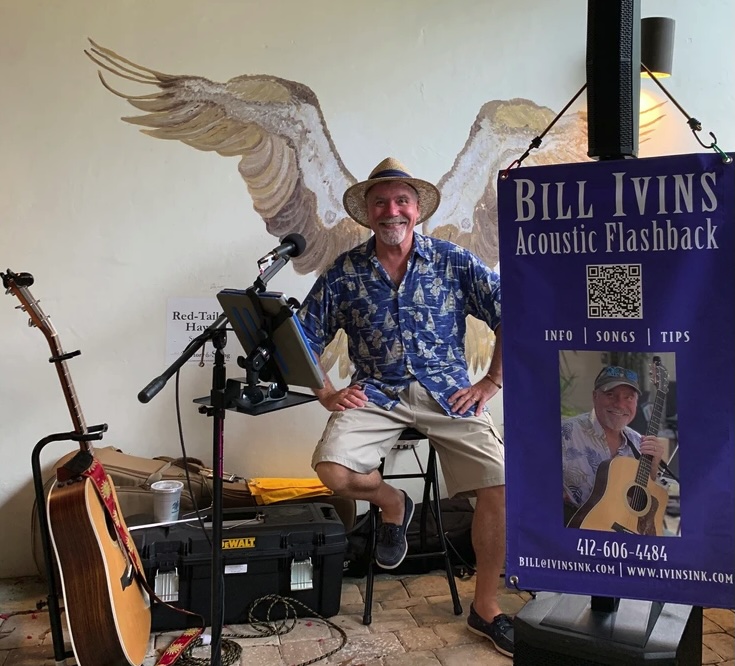 A smiling man in a blue shirt and straw hat sits in front of a microphone and music setup, with a guitar beside him and painted bird wings on the wall behind. A banner for Bill Ivins Acoustic Flashback is displayed on the right.