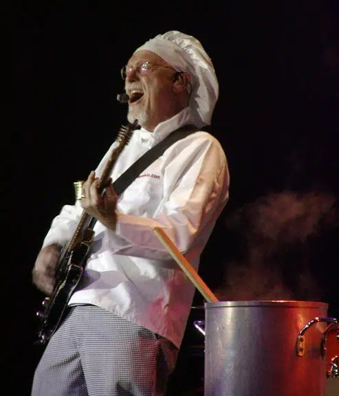 A musician dressed as a chef, wearing a white chef hat and coat, plays an electric guitar next to a steaming pot with a large wooden spoon on stage.