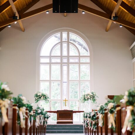 A bright, empty church interior with wooden pews decorated with white flowers and ribbons, leading to an altar adorned with floral arrangements and a gold cross, beneath a large arched window.
