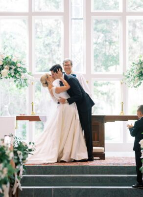 A bride and groom share a kiss at the altar during their wedding ceremony, with a smiling officiant behind them and a young boy clapping nearby. The scene is set in a bright, floral-decorated chapel.