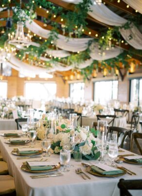 An elegant wedding reception hall with long tables set with white linens, green and white floral centerpieces, glassware, and plates. The ceiling is decorated with greenery, draped fabric, and string lights.